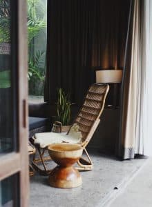 Sedia in stile naturale con cuscino e pianta decorativa, in un ambiente interno con tende e illumina.