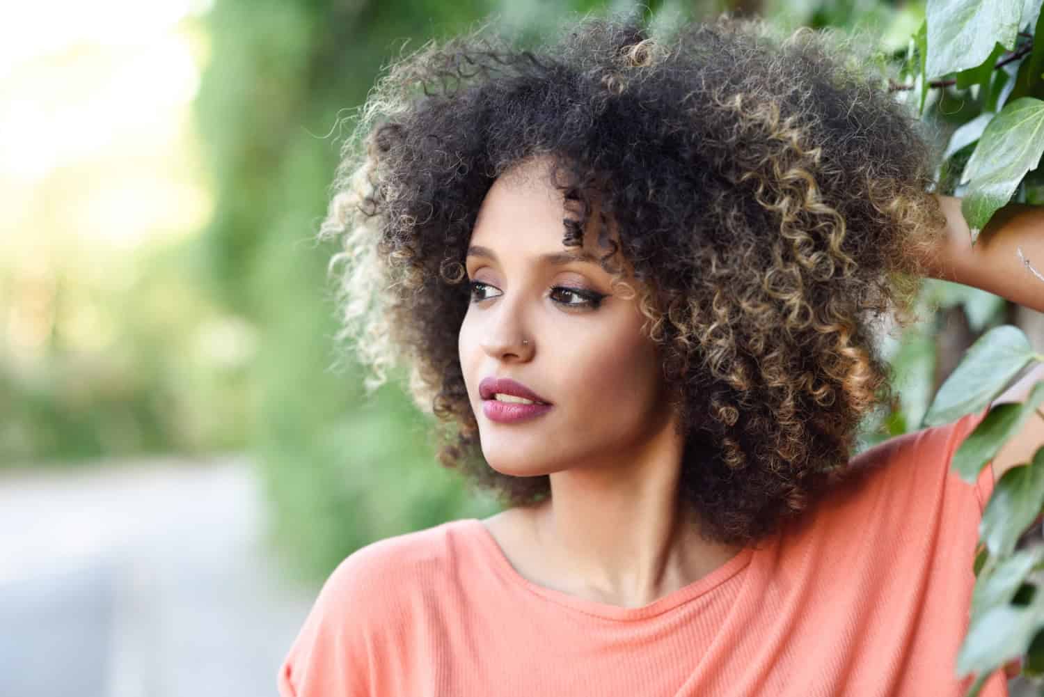 Donna con capelli ricci naturali che si appoggia alle foglie verdi in un ambiente all'aperto.