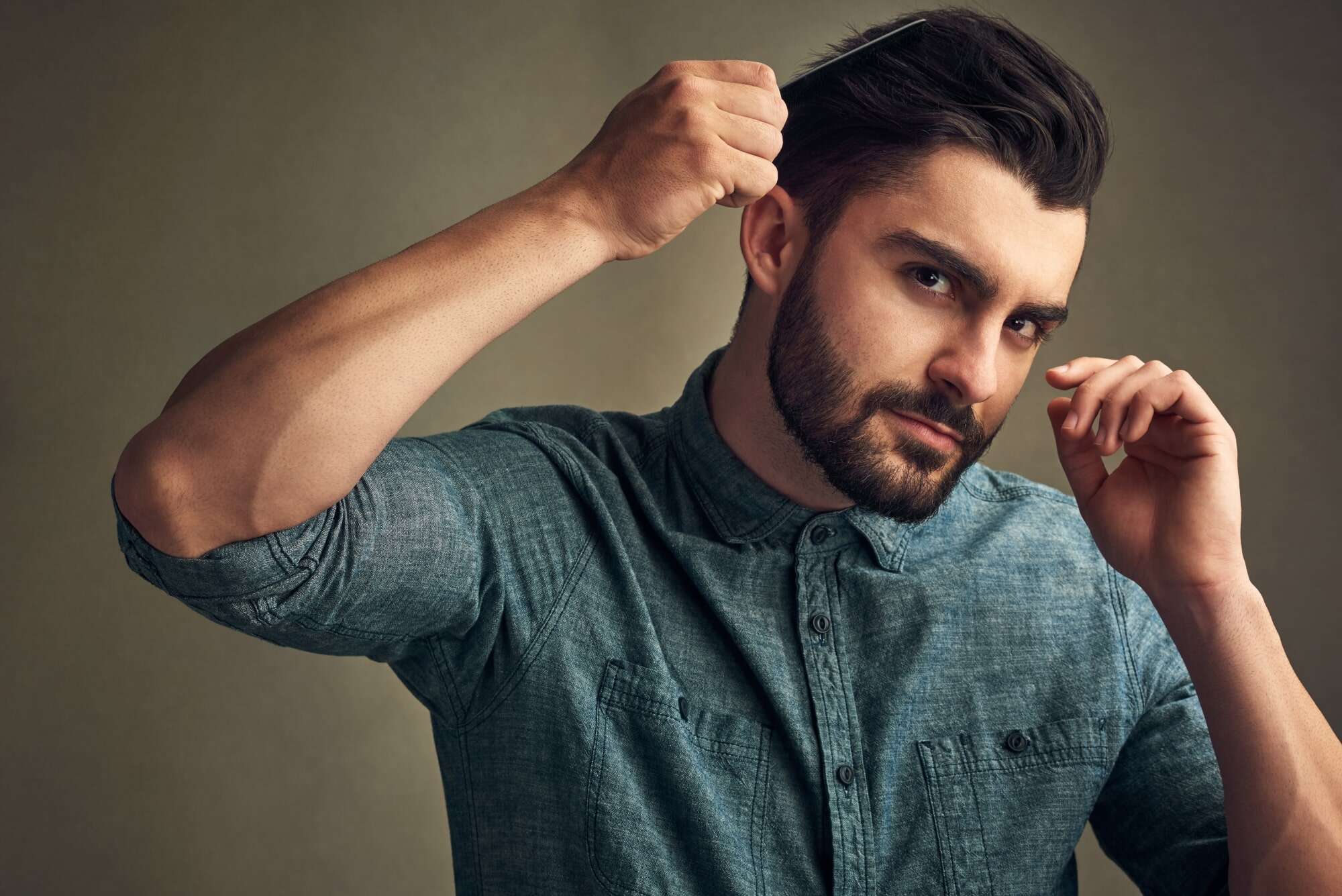 Uomo con barba che si sistema i capelli, indossa una camicia blu scuro, sfondo neutro.